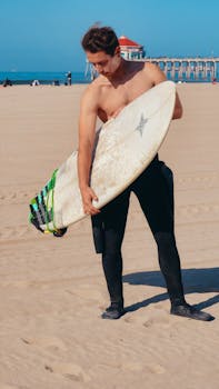 A young male surfer holding his surfboard on a sunny beach with a pier in the background, ready to hit the waves.