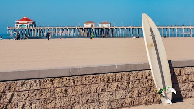 Sunny day at Huntington Beach with a surfboard resting on a brick wall and iconic pier in the background.