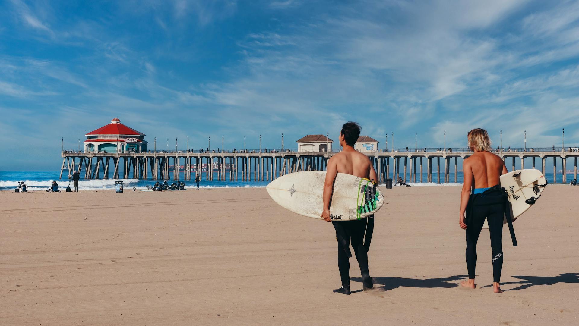 Two surfers at Huntington Beach, California, gaze at the ocean near the iconic pier on a sunny day.