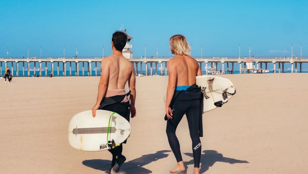 Two surfers carrying surfboards on the beach near a sunny pier.