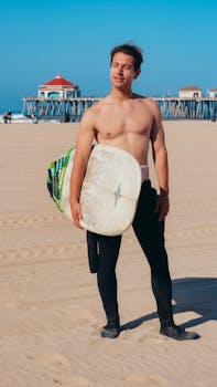 Man with surfboard on the sandy beach by pier, showcasing a sunny California day.