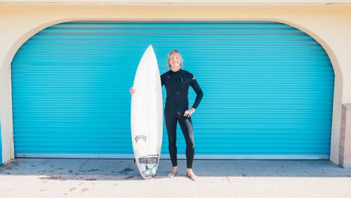 Free A surfer in a wetsuit poses with a surfboard against a vibrant blue roller shutter. Stock Photo