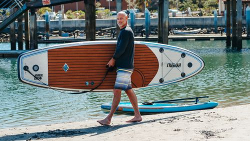 Free Adult man walking on beach carrying a paddleboard. Perfect for outdoor lifestyle and adventure themes. Stock Photo