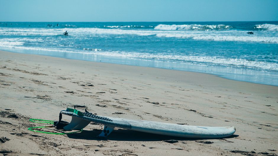 Vacant surfboard rests on a sandy beach with ocean waves and surfers in the distance.