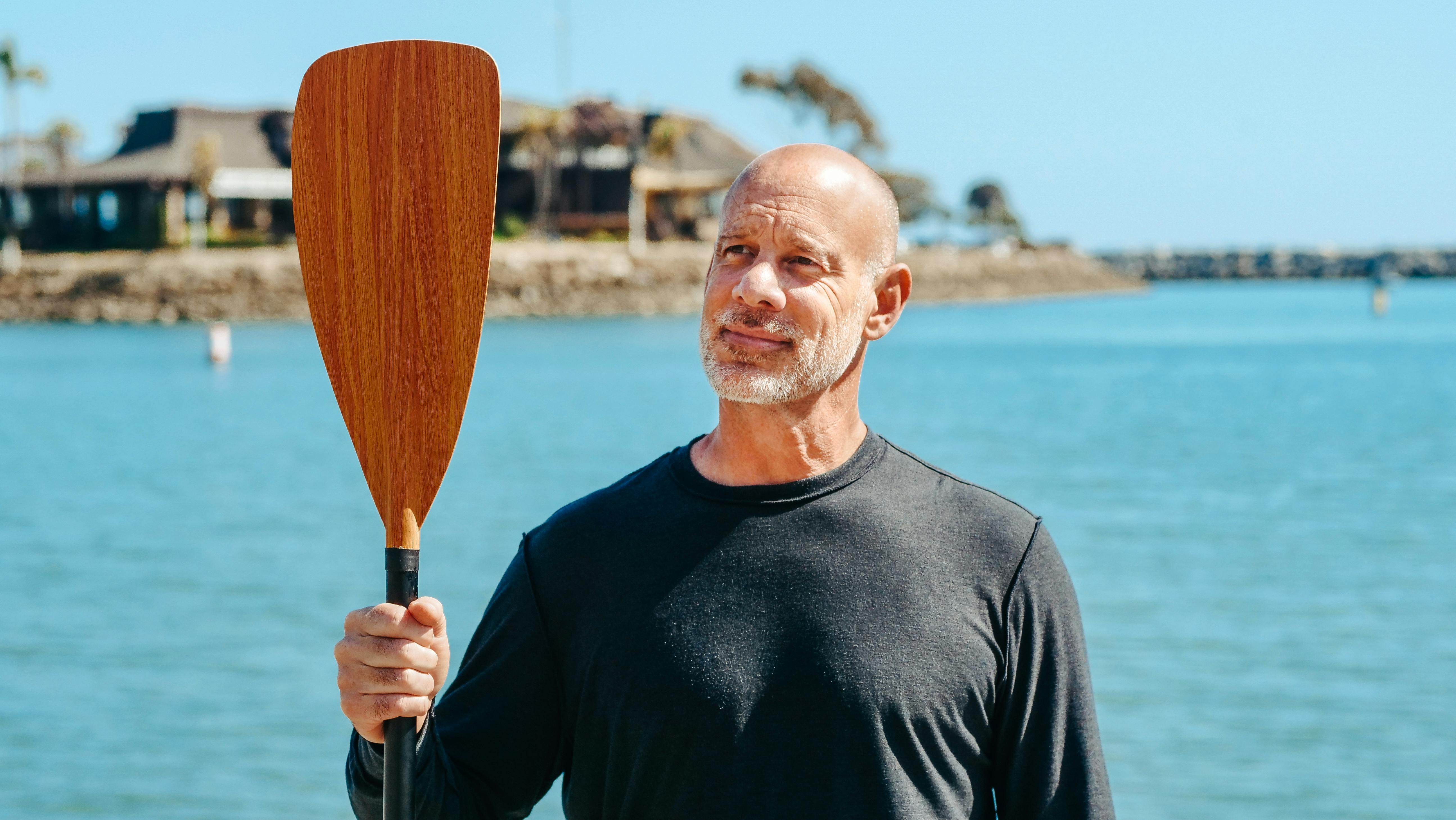 Elderly man holding paddle by the lake on a sunny day, enjoying relaxation and recreation.