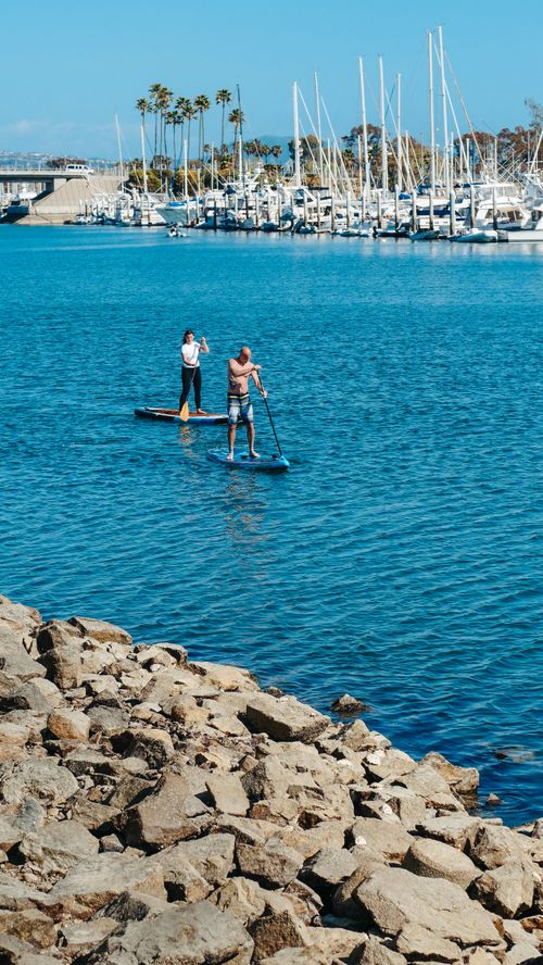 Free Two adults paddleboarding near a marina on a sunny summer day, enjoying outdoor recreation. Stock Photo