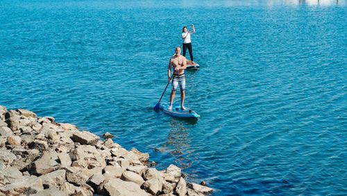 Free Two adults paddle boarding on a tranquil ocean by a rocky shore on a sunny day. Stock Photo