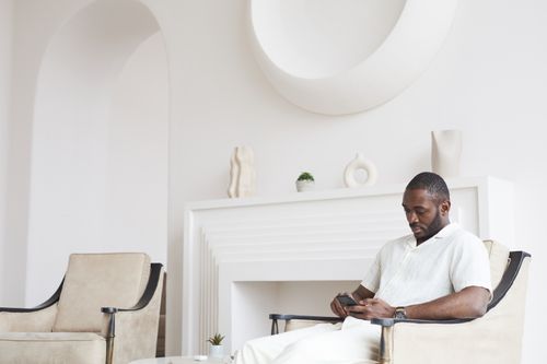 Free African American man sitting in a modern white room using a smartphone. Stock Photo