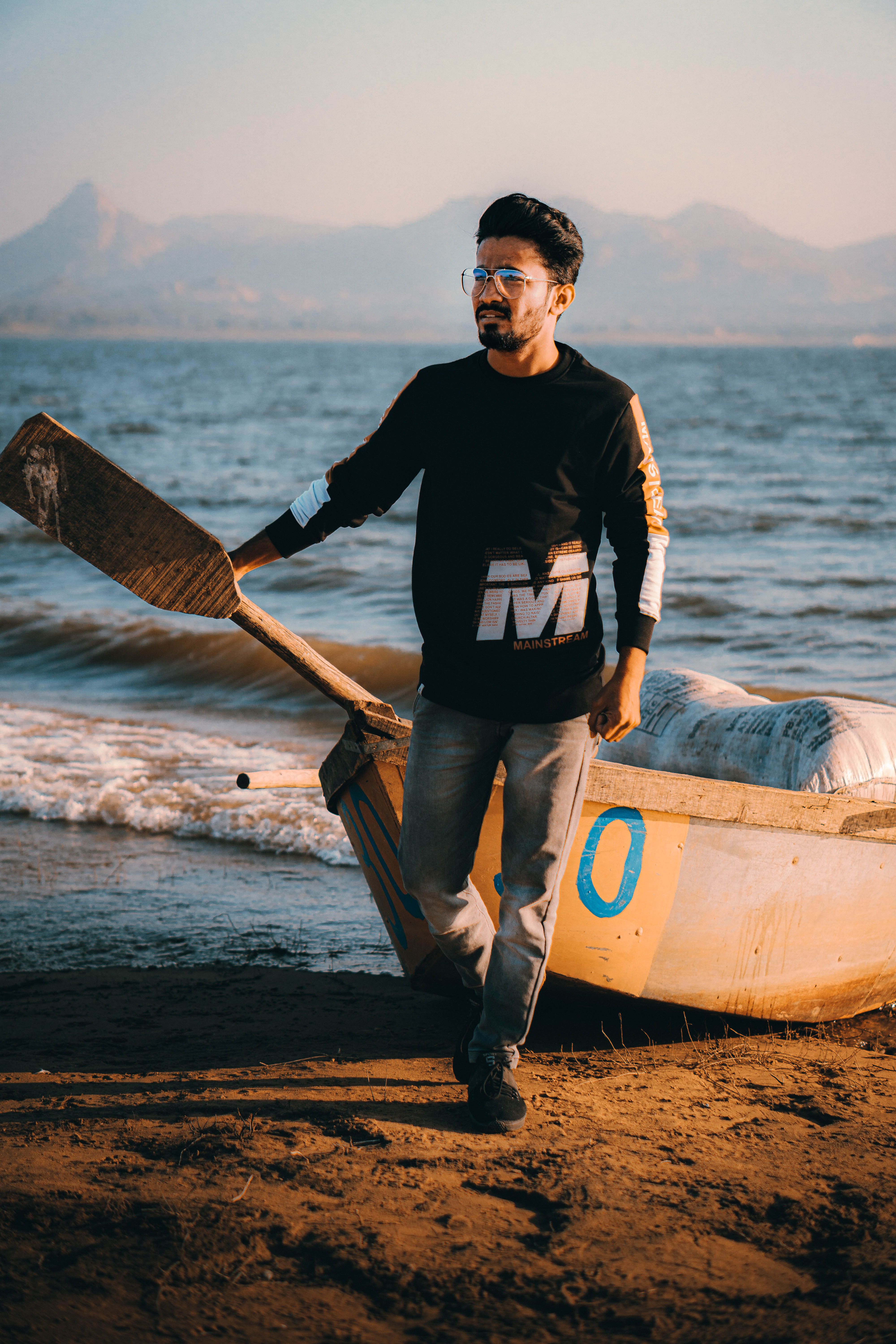 A fashionable man in sunglasses poses by a beached boat with mountains in the background.