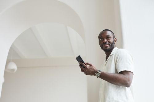 Free Black man in a light shirt using a smartphone, standing indoors with a bright, modern background. Stock Photo