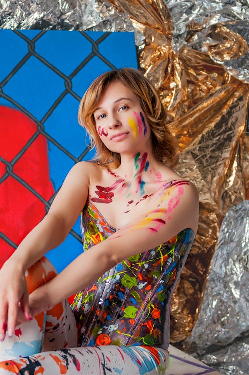 Portrait of young red haired young woman with painting and gold and silver foil on background wearing corset painted with vivid colors while looking at camera