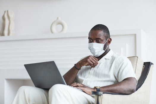 African American man working from home while wearing a face mask, utilizing a laptop. Indoor setting.