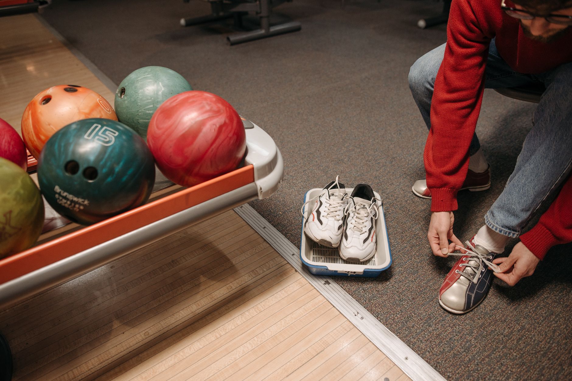 A man ties rental bowling shoes beside colorful bowling balls in an alley.