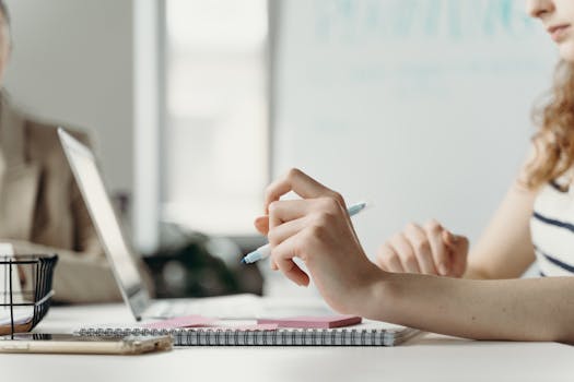 Close-up of a woman in an office, planning on a laptop with notes and pen.