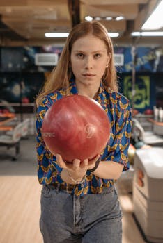 A young woman in a bowling alley holding a red bowling ball, preparing for a game.