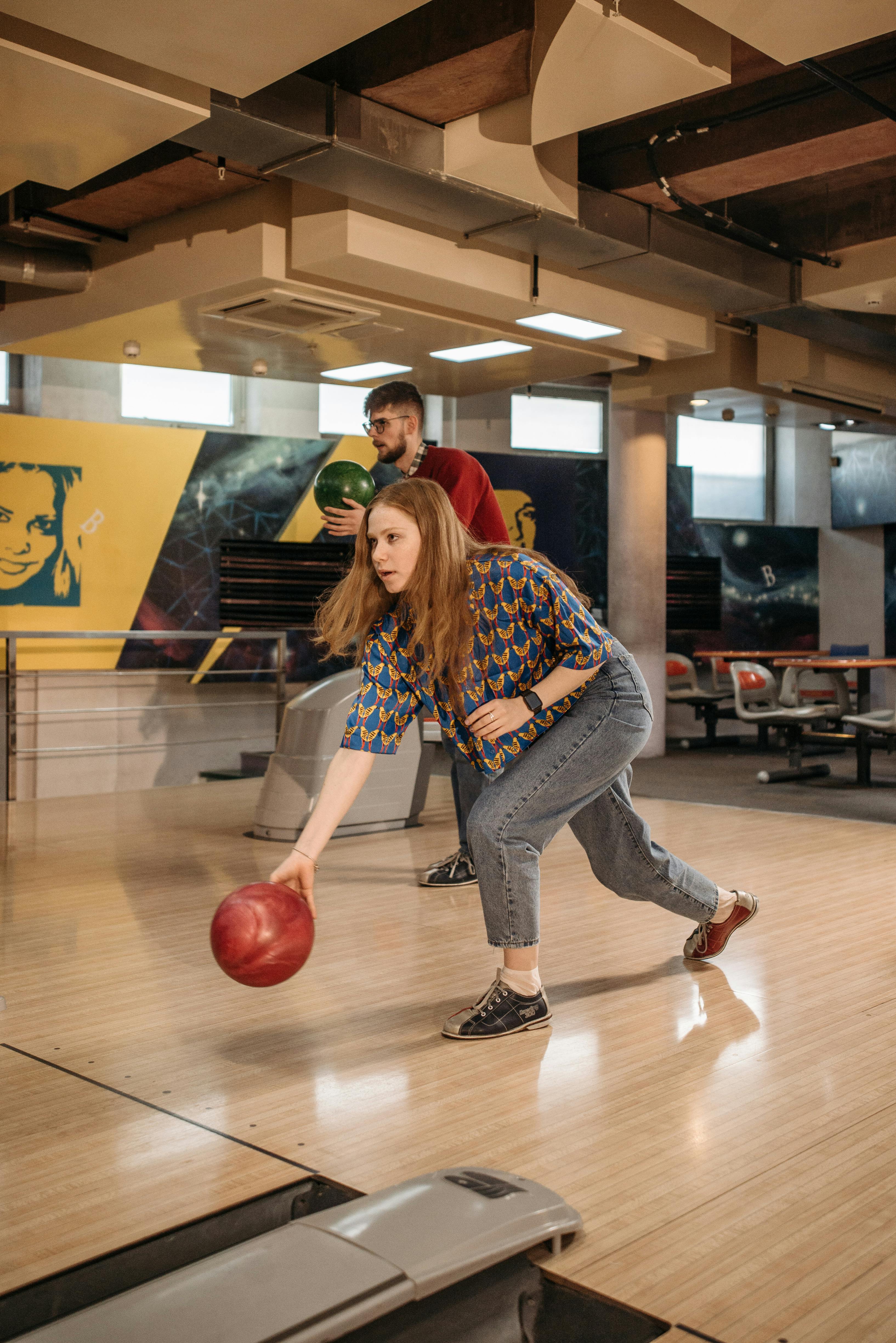 Woman bowling, red ball in motion, man in background. Bowling alley setting, wooden floor, colorful mural.