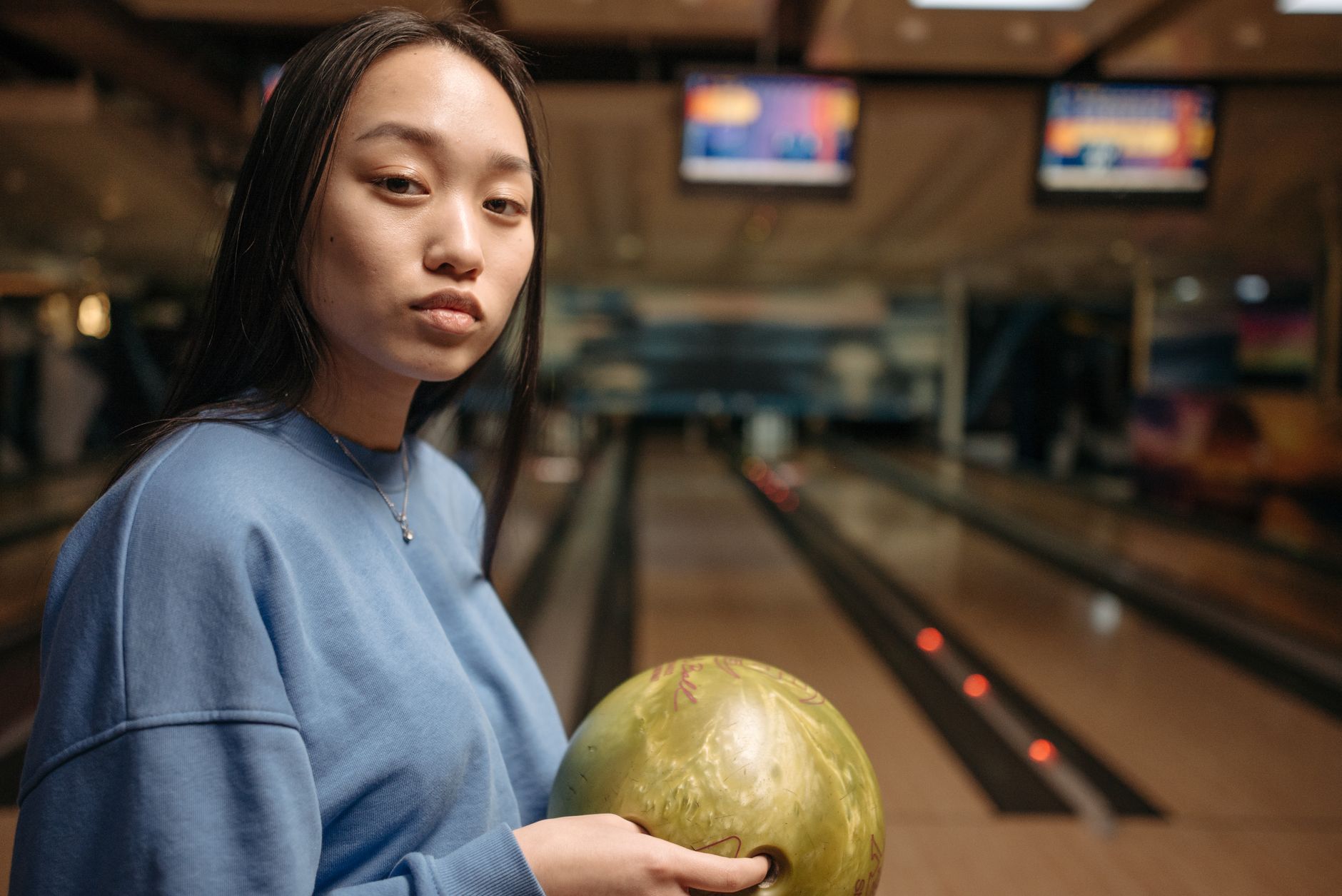 East Asian woman holding a green bowling ball in a bowling alley, ready to play.