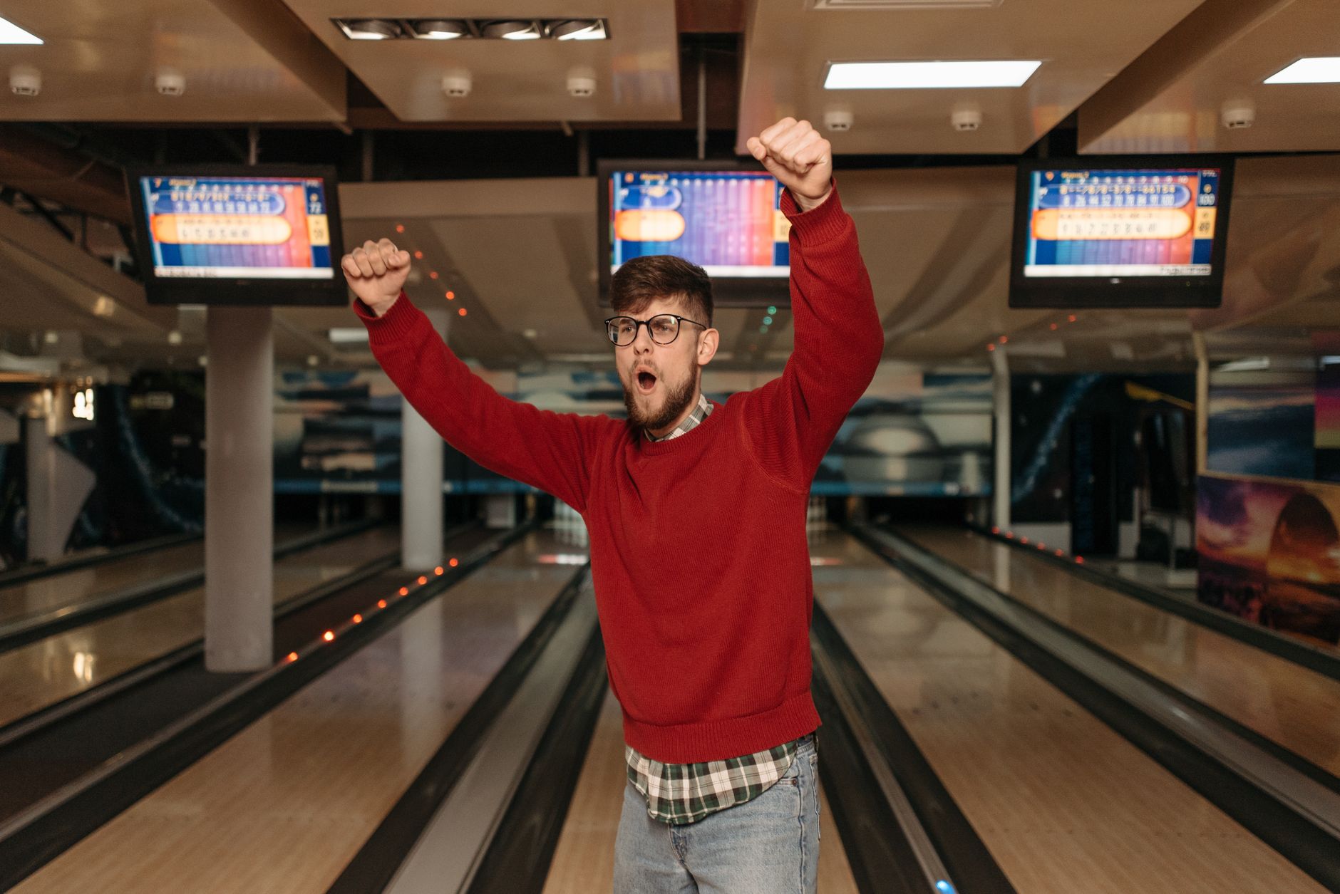 A man in a red sweater celebrates a successful bowling strike indoors.