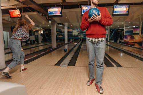 Man in Re Sweater Holding Blue Ball Standing Beside a Woman Playing Bowling