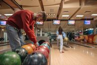 A man in a red sweater selects a bowling ball while a woman rolls a ball down the lane.