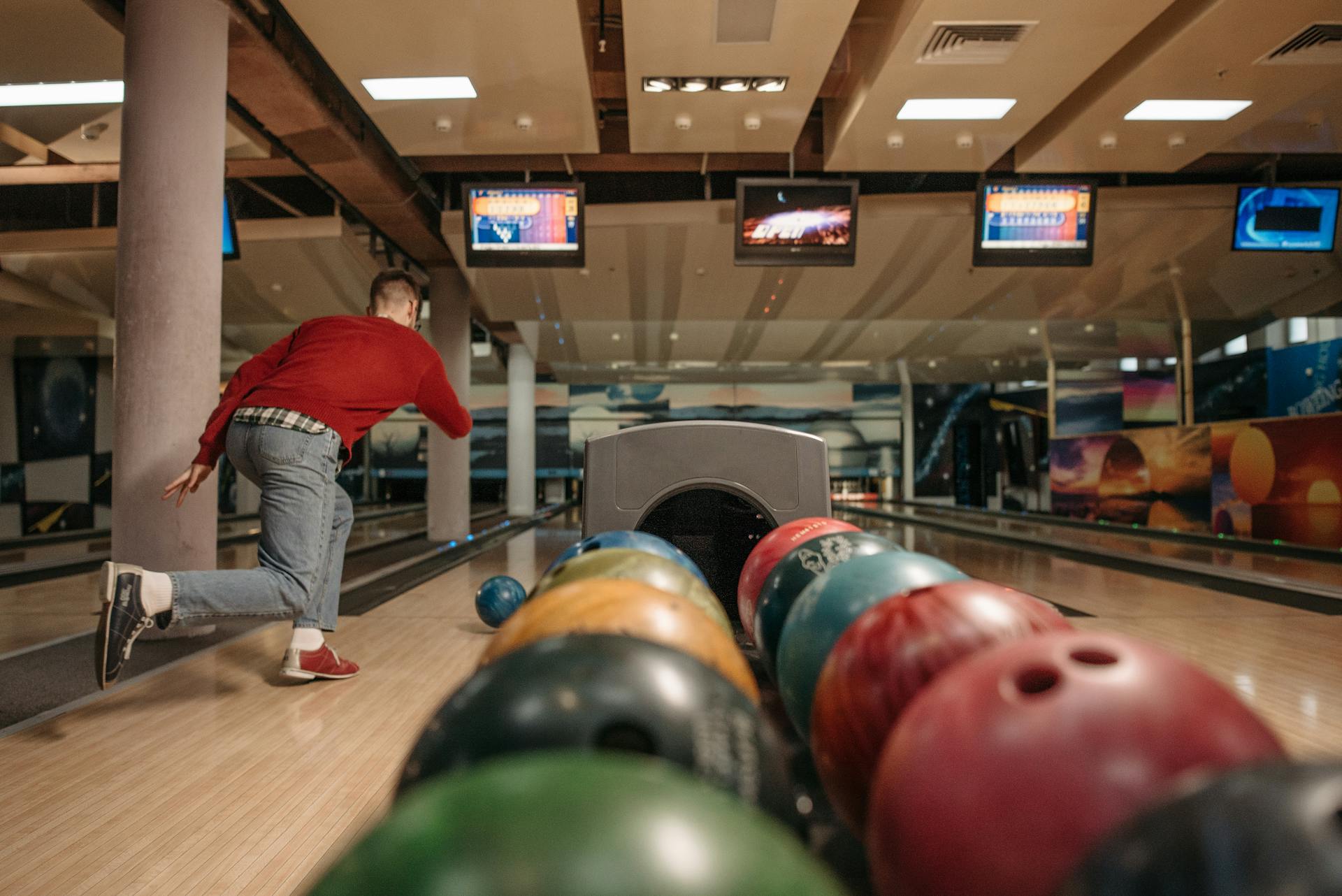 A man in a bowling alley throwing a bowling ball down a lane with colorful balls in the foreground.