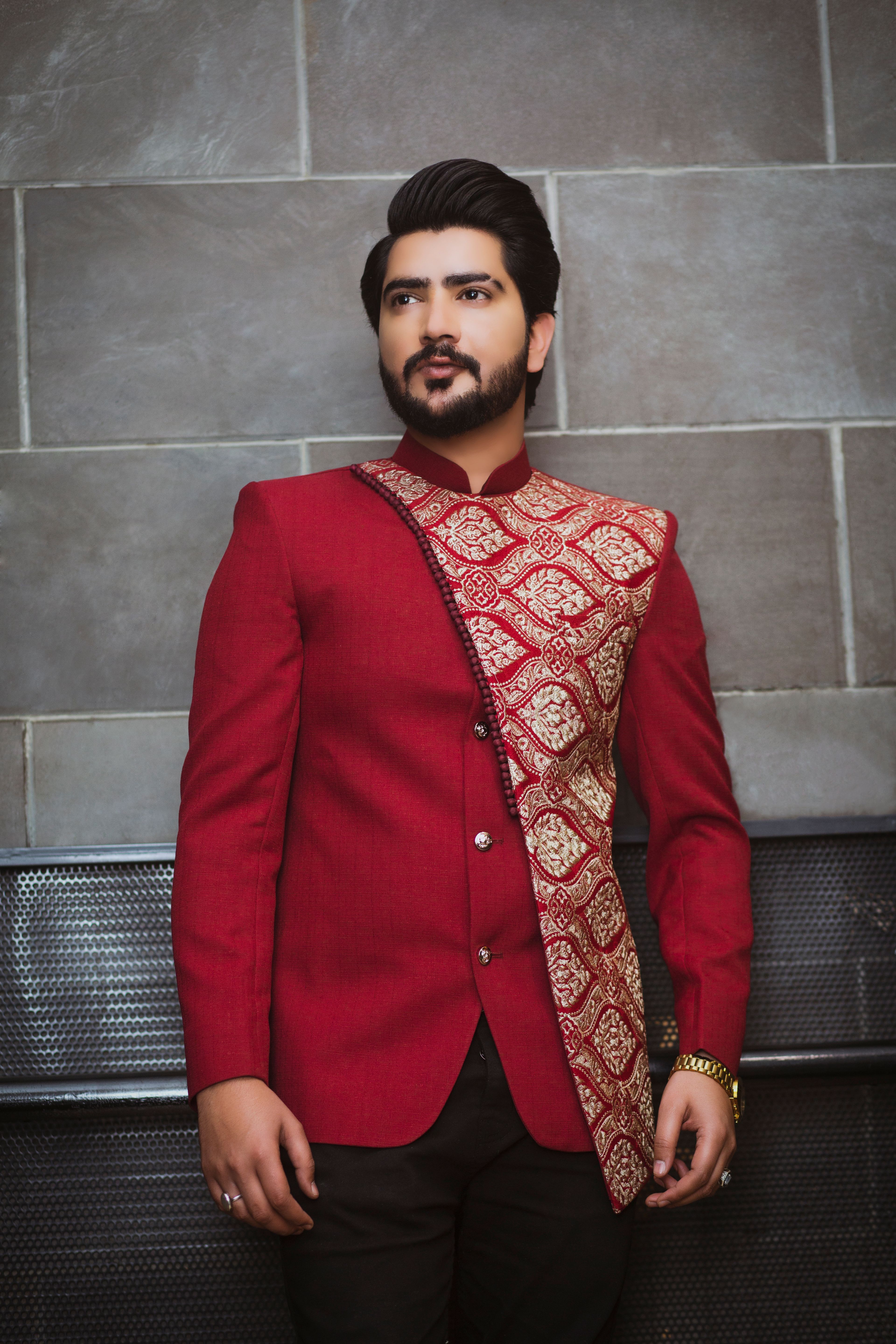 Man in a red traditional outfit with ornate patterns posing against a textured wall.