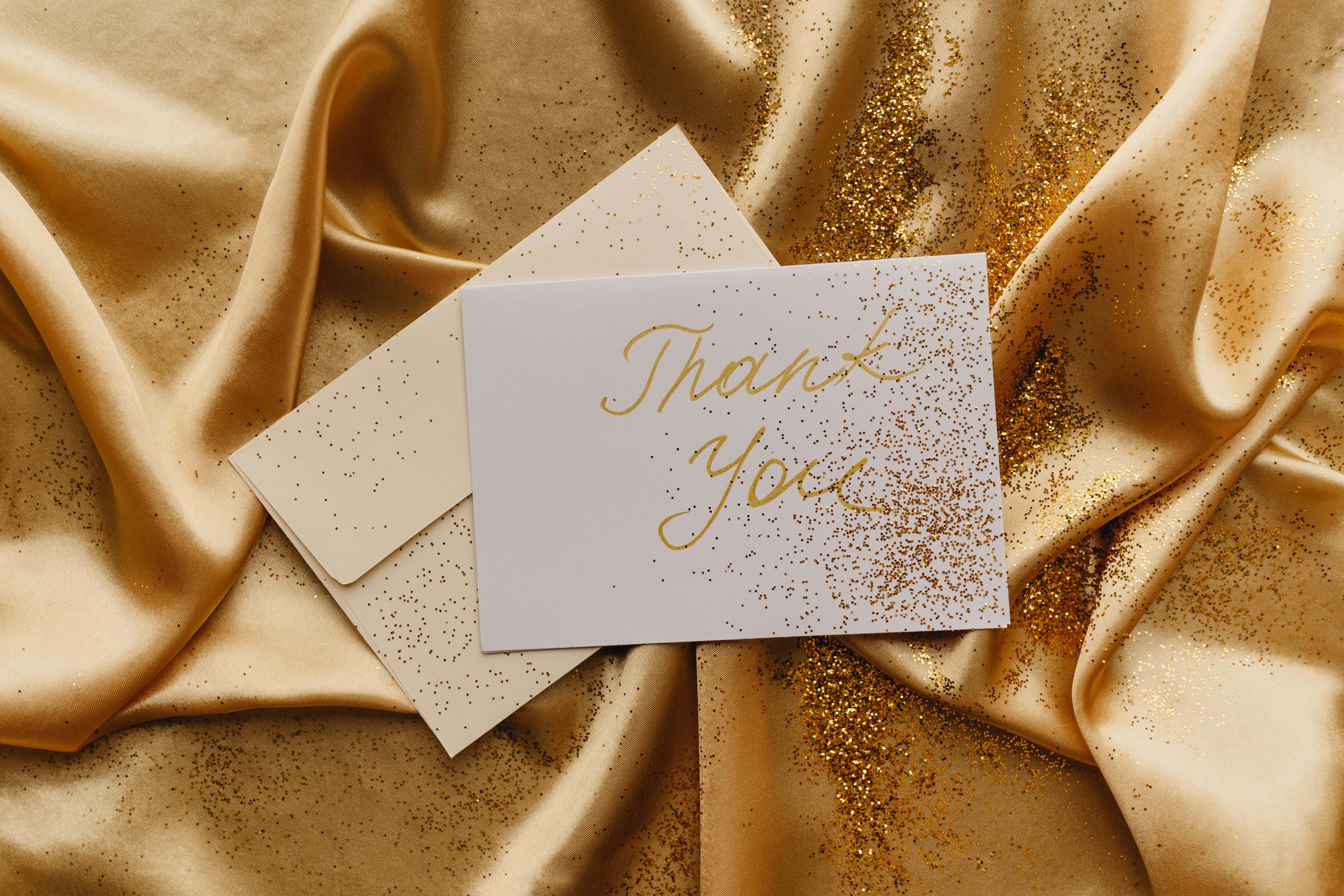Elegant thank you card displayed on gold satin with scattered glitter.