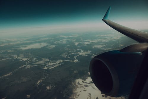 Through window of modern wing and turbine of airplane flying in sky above verdant land