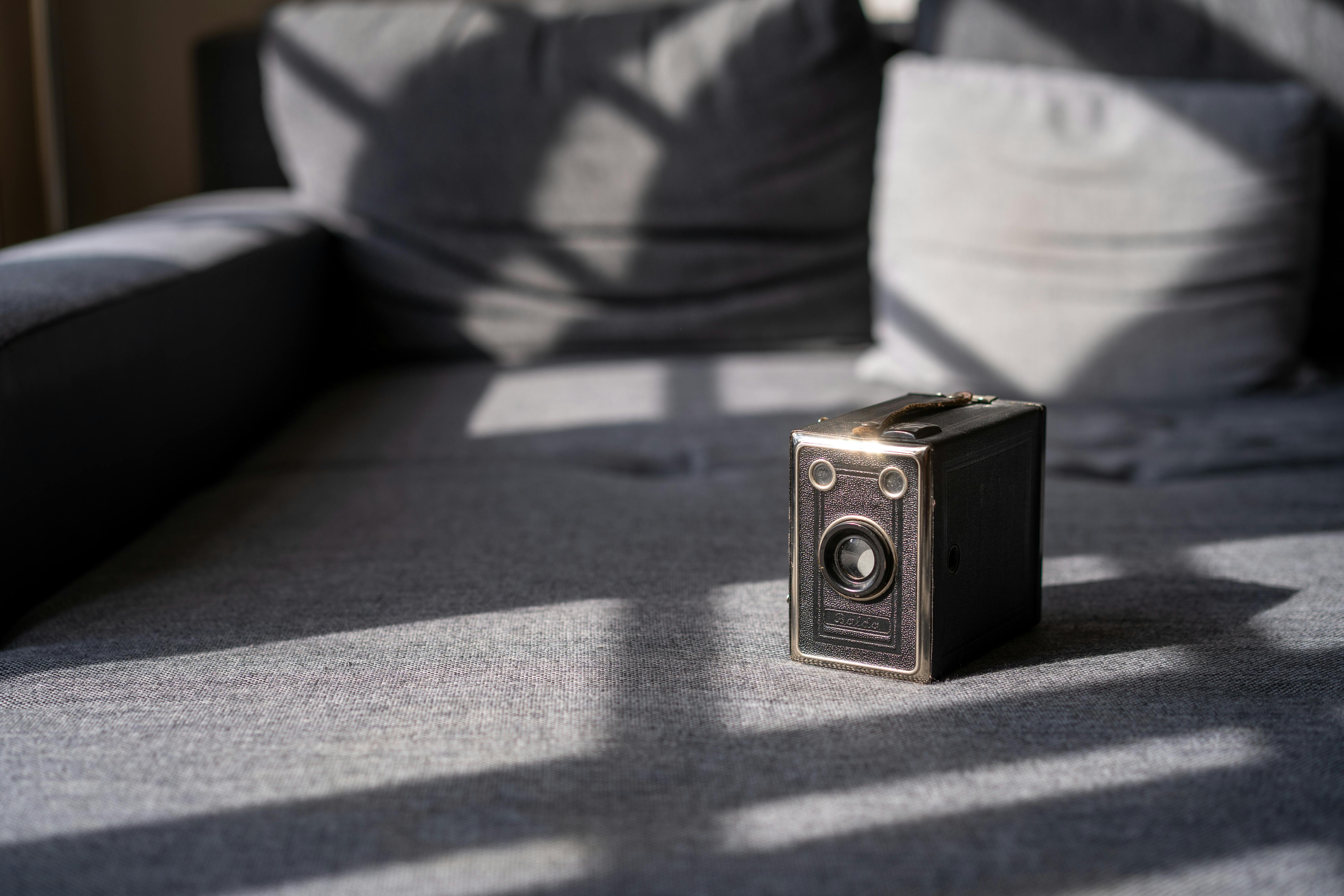 A classic vintage box camera rests on a gray couch, beautifully lit by natural sunlight creating artistic shadows.
