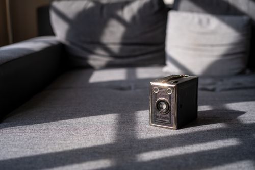 Free A classic vintage box camera rests on a gray couch, beautifully lit by natural sunlight creating artistic shadows. Stock Photo