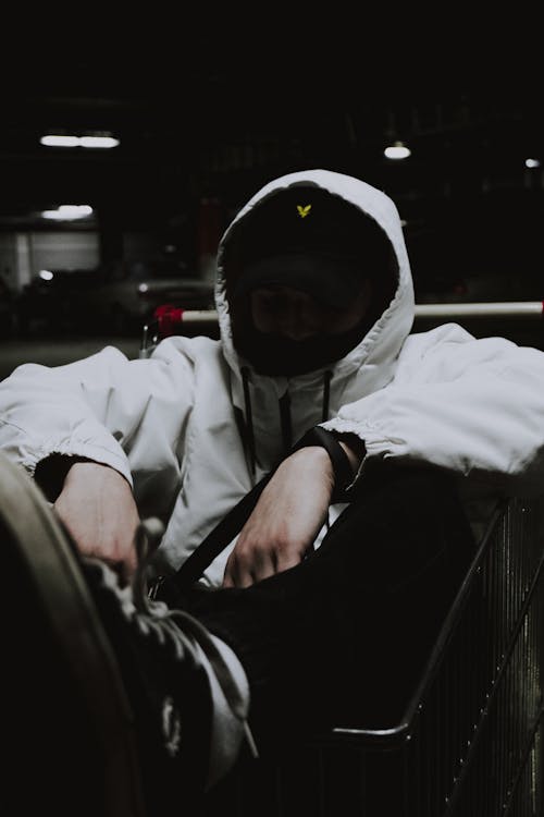 Anonymous person in hoodie and sneakers with black mask sitting in shopping cart at night