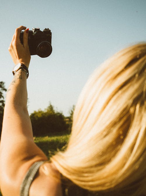 Back view of crop anonymous female traveler with long blond hair taking selfie on modern photo camera while standing in grassy field against cloudless blue sky on sunny day