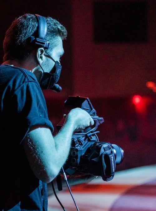 Free Man filming with camera indoors wearing a mask, focused on capturing quality footage. Stock Photo