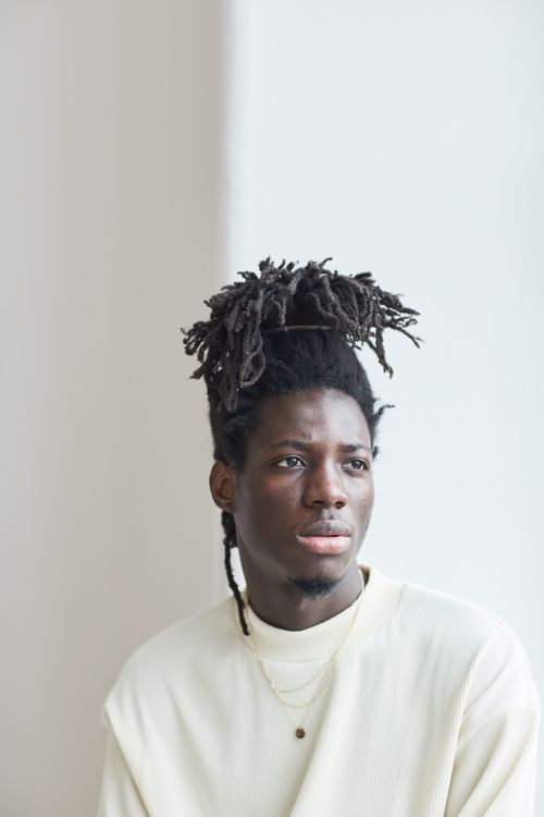Free Pensive black man with dreadlocks wearing a white sweater looks away in a calm indoor setting. Stock Photo