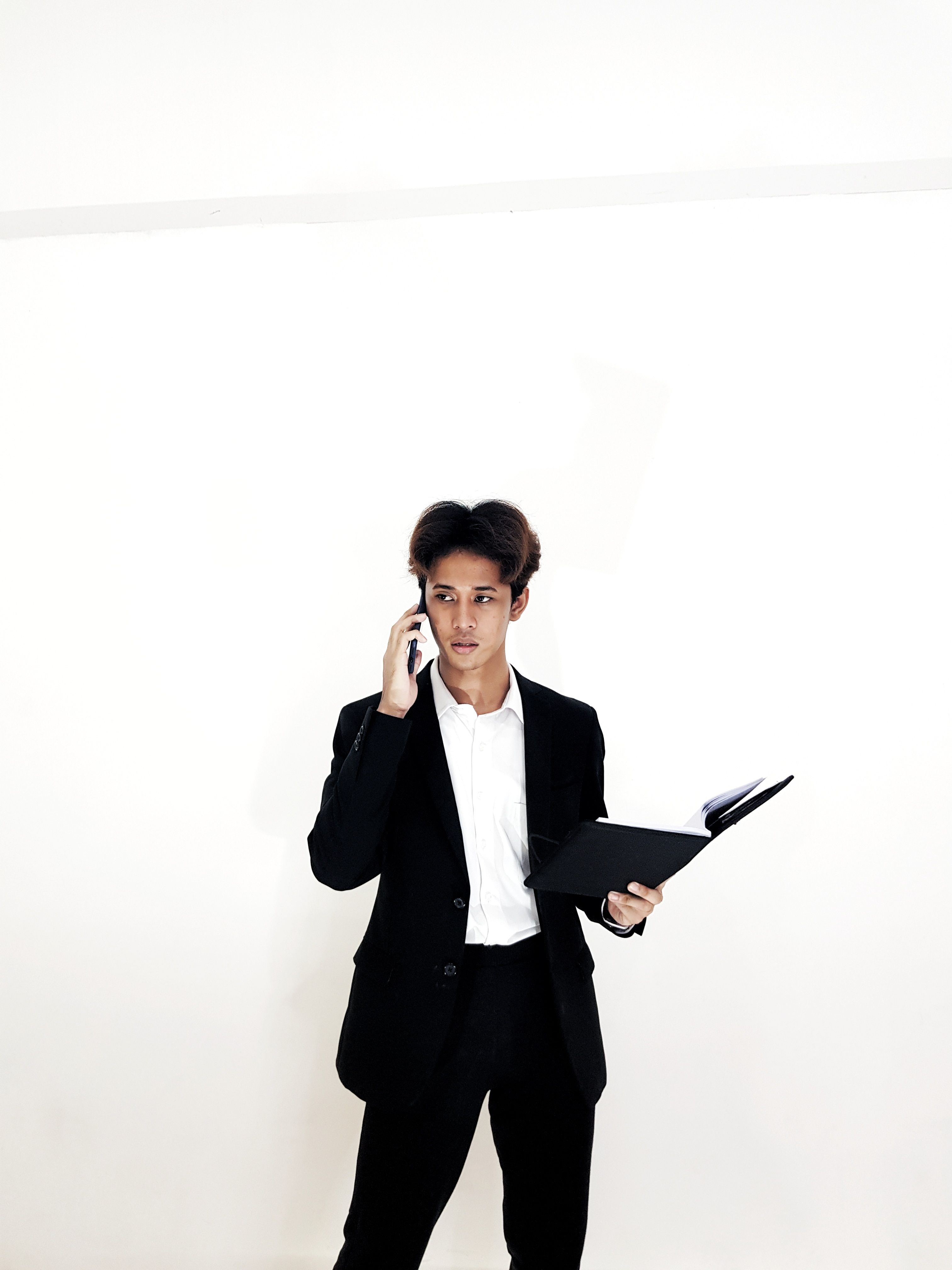 Asian businessman in a suit talking on a smartphone and holding a notebook, standing against a plain background. - Fotografer Sidoarjo