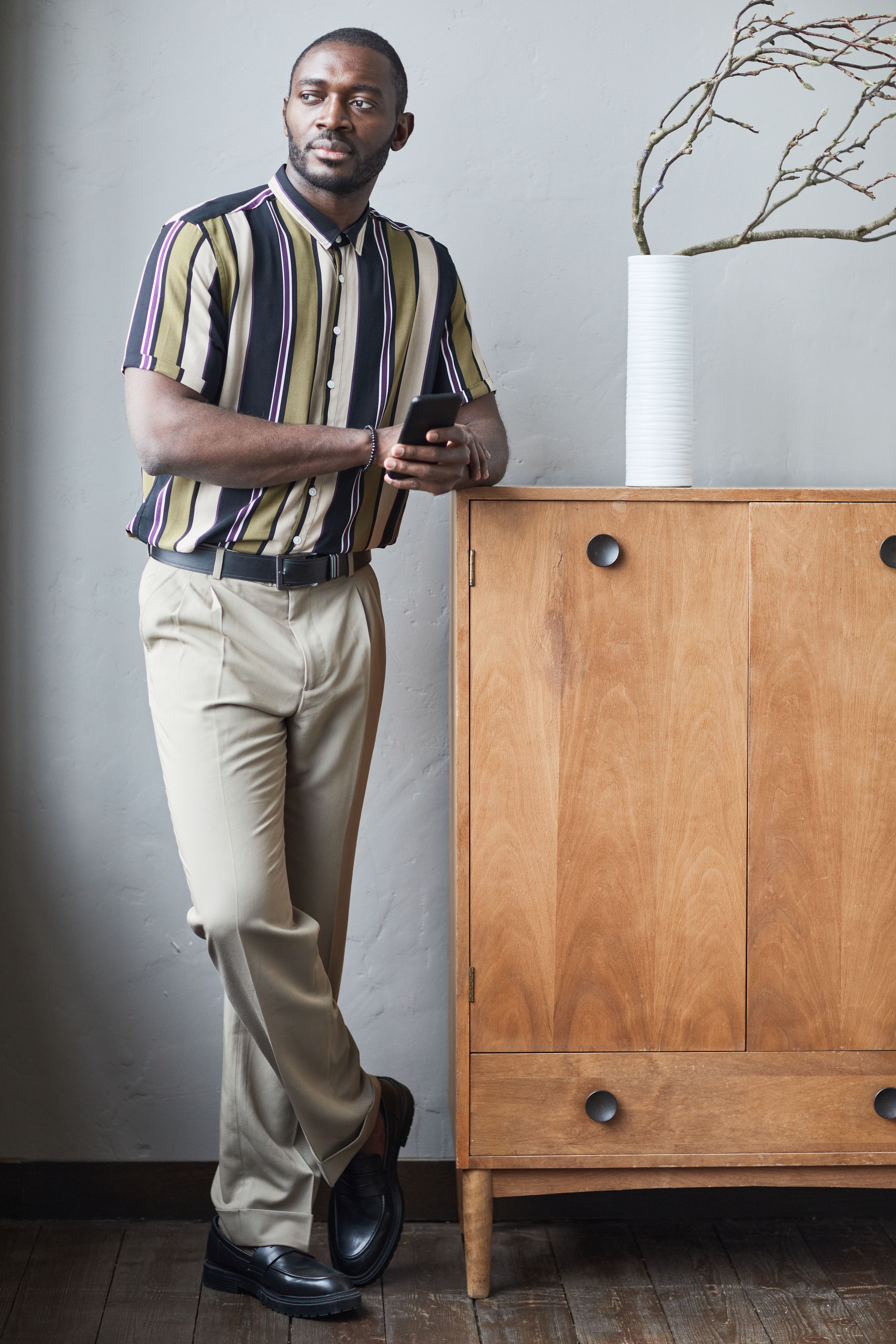 Stylish man in striped shirt leaning against a wooden cabinet, holding a smartphone.