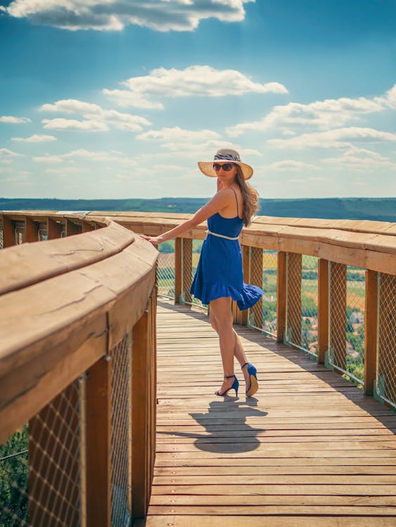 A Woman in Blue Dress Wearing a Hat and Sunglasses while Standing on a Wooden Bridge