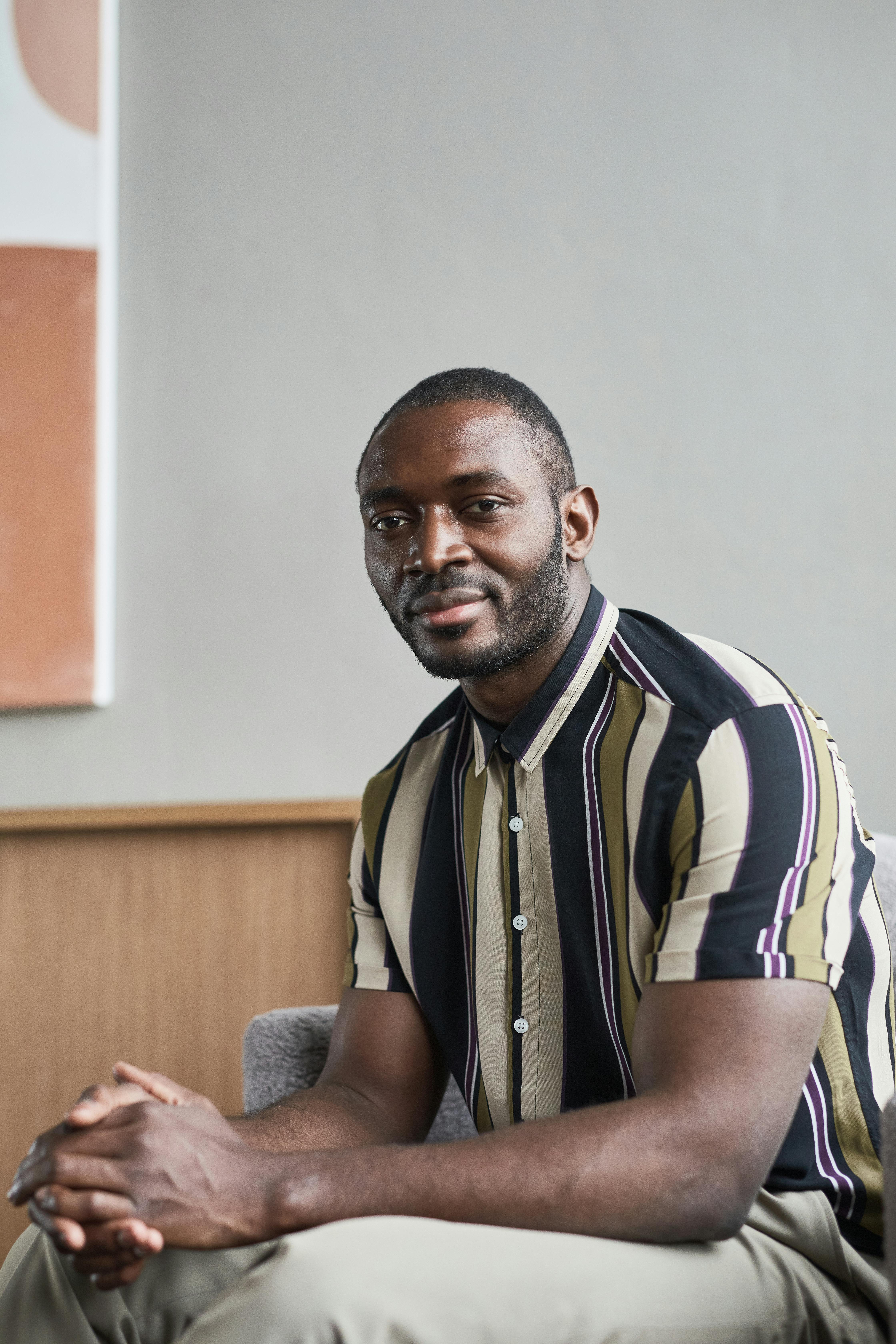 A handsome bearded man in a striped shirt smiling while seated in a modern living room.