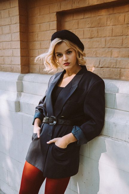 Serious young female in beret and trendy outfit leaning on building wall in town street in daytime while looking at camera
