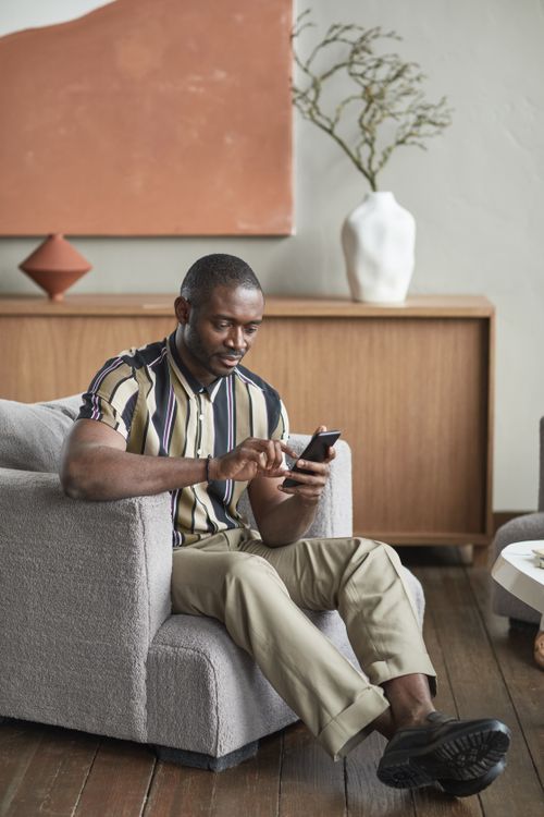 Free Casually dressed man sitting in armchair using smartphone in modern living room. Stock Photo