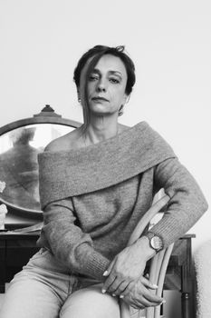 Stylish woman wearing an off-shoulder sweater poses indoors by a mirror in grayscale.