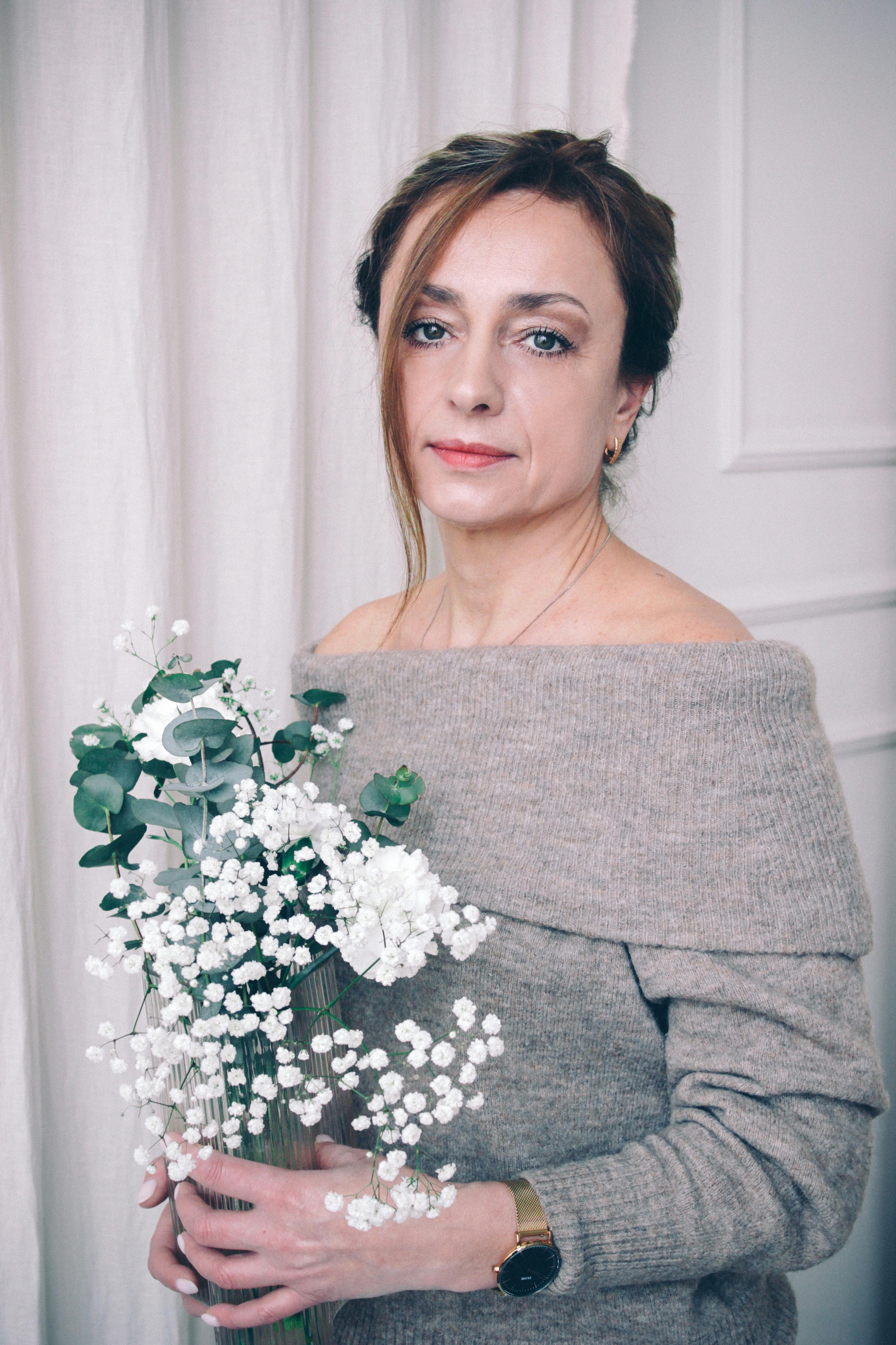 A woman in a cozy off-shoulder sweater holds a bouquet of white flowers, creating a serene indoor ambiance.
