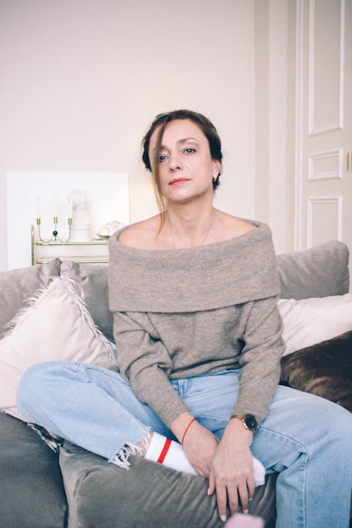 A Woman in Gray Off Shoulder Top and Denim Jeans Sitting on the Couch