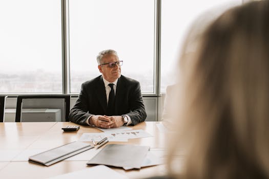 Senior businessman in a corporate meeting, wearing a black suit and glasses, indoors.