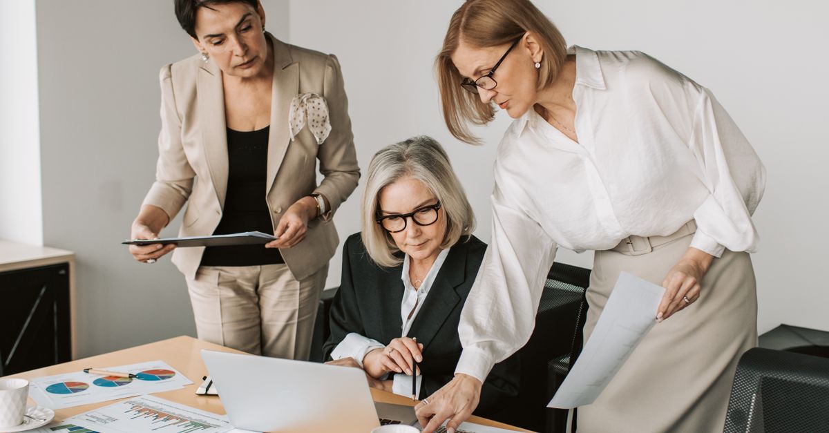 Three senior businesswomen in an office working together on a laptop and documents.