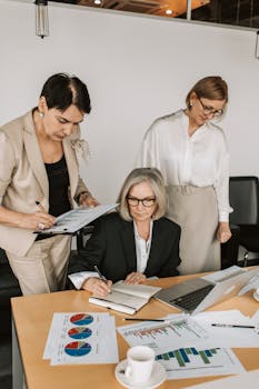 Three women in business attire work together on financial reports at an office table.