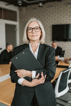 Senior businesswoman in formal attire holding a clipboard during a meeting in an office setting.