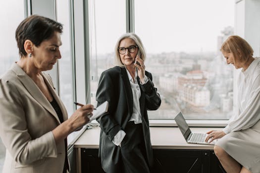 Three professional women engaging in office work with laptops and notebooks by a window.
