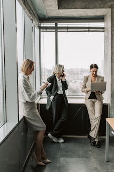 Three businesswomen in a modern office setting, working with a laptop and taking notes.