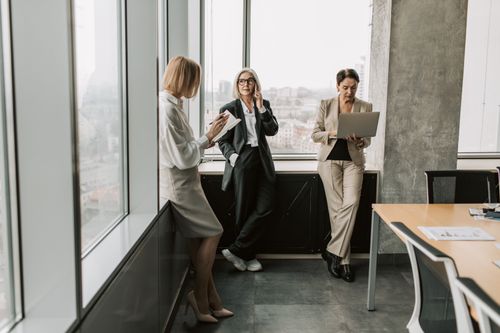 Free Three women in office attire collaborating near large windows, using technology. Stock Photo